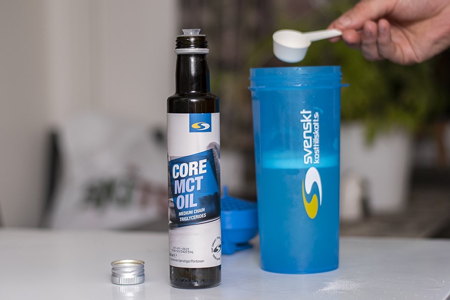 Billede af Core MCT-olie i flaske, en shaker i baggrunden og en hånd, der holder en 1 spsk MCT-olie i shakeren.