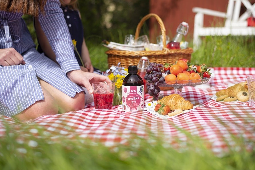 Picknick-filt med frukt, saft och en flaska Natusaft C+D, med mamma och dotter i bakgrunden.